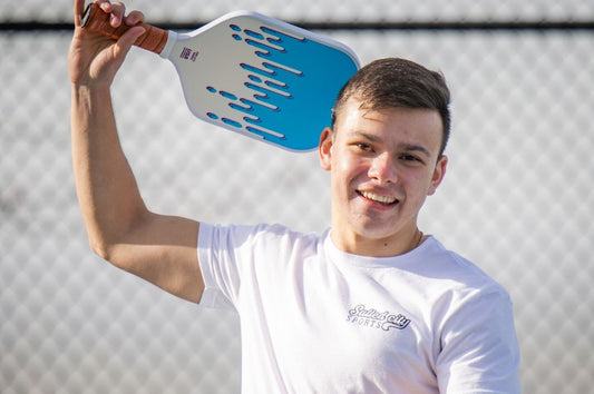 A man holding a pickleboard paddle in front of a fence, preparing for a pickleball warm-up routine.