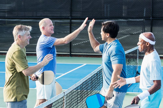 Four men high five on a tennis court, celebrating the senior pickleball benefits of teamwork and physical activity.