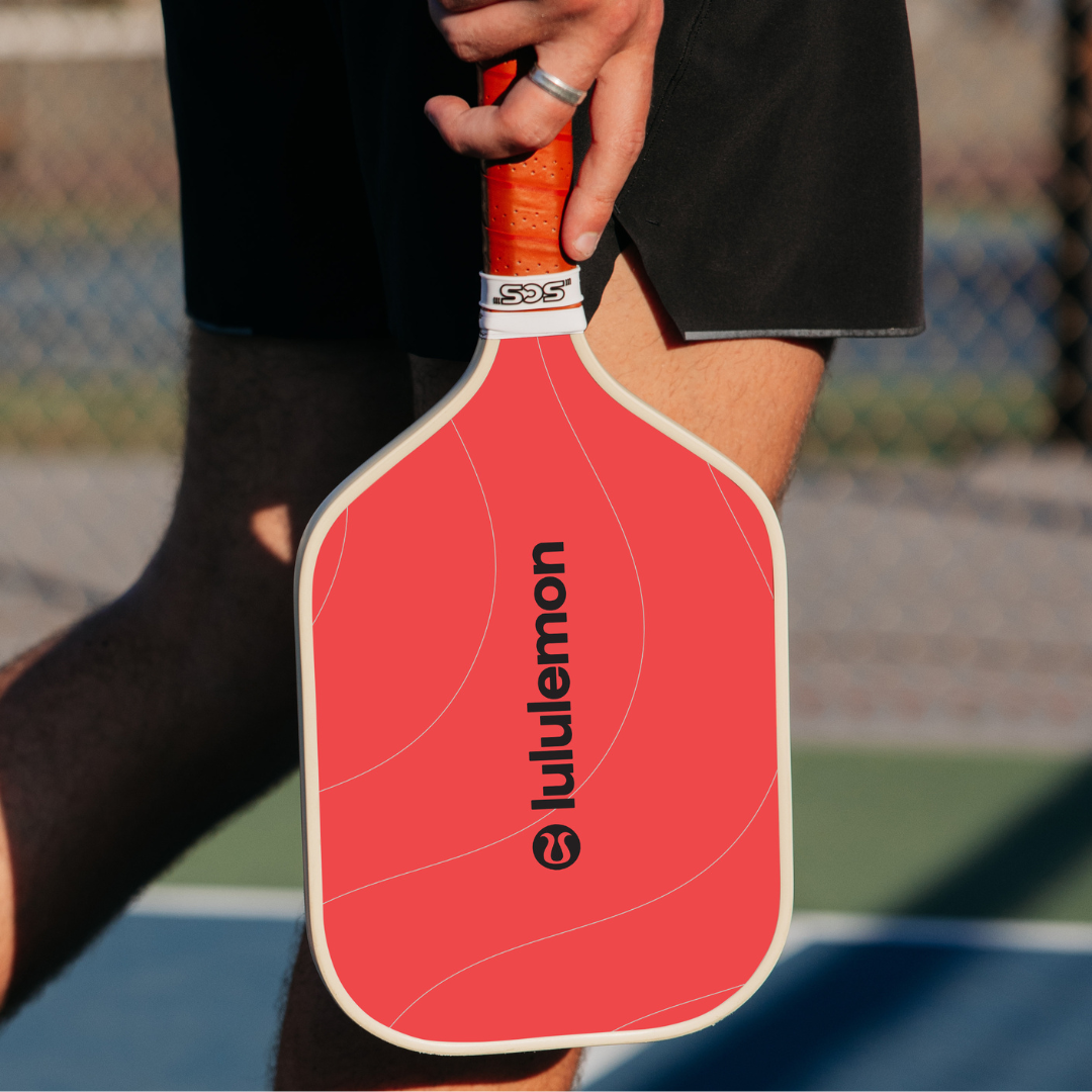 Person holding a red pickleball paddle with 'lululemon' branding on a blurred court background