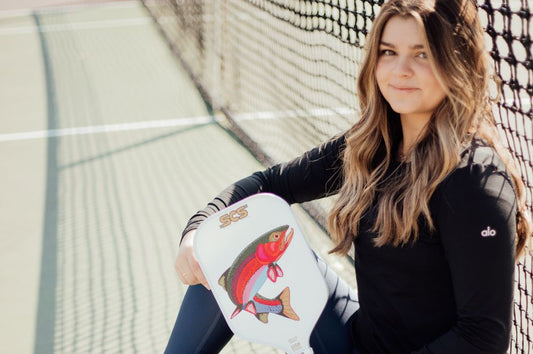 A girl sitting on a tennis court, holding a custom pickleball paddle beside her.