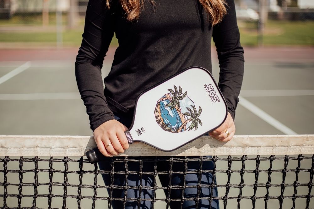 A young woman standing on a tennis court holding a new custom picklebal paddle design by Salted City Sports