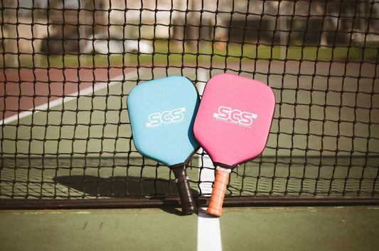 A blue and pink pickleball paddle rest against a net showcasing youth pickleball equipment.