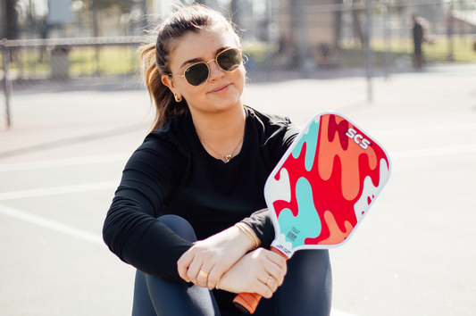 A woman sitting on the ground, thoughtfully choosing the best pickleball paddle for her game.