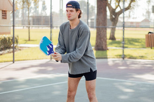 A man in shorts and a cap holds a tennis racket, preparing for pickleball drills on a sunny court.