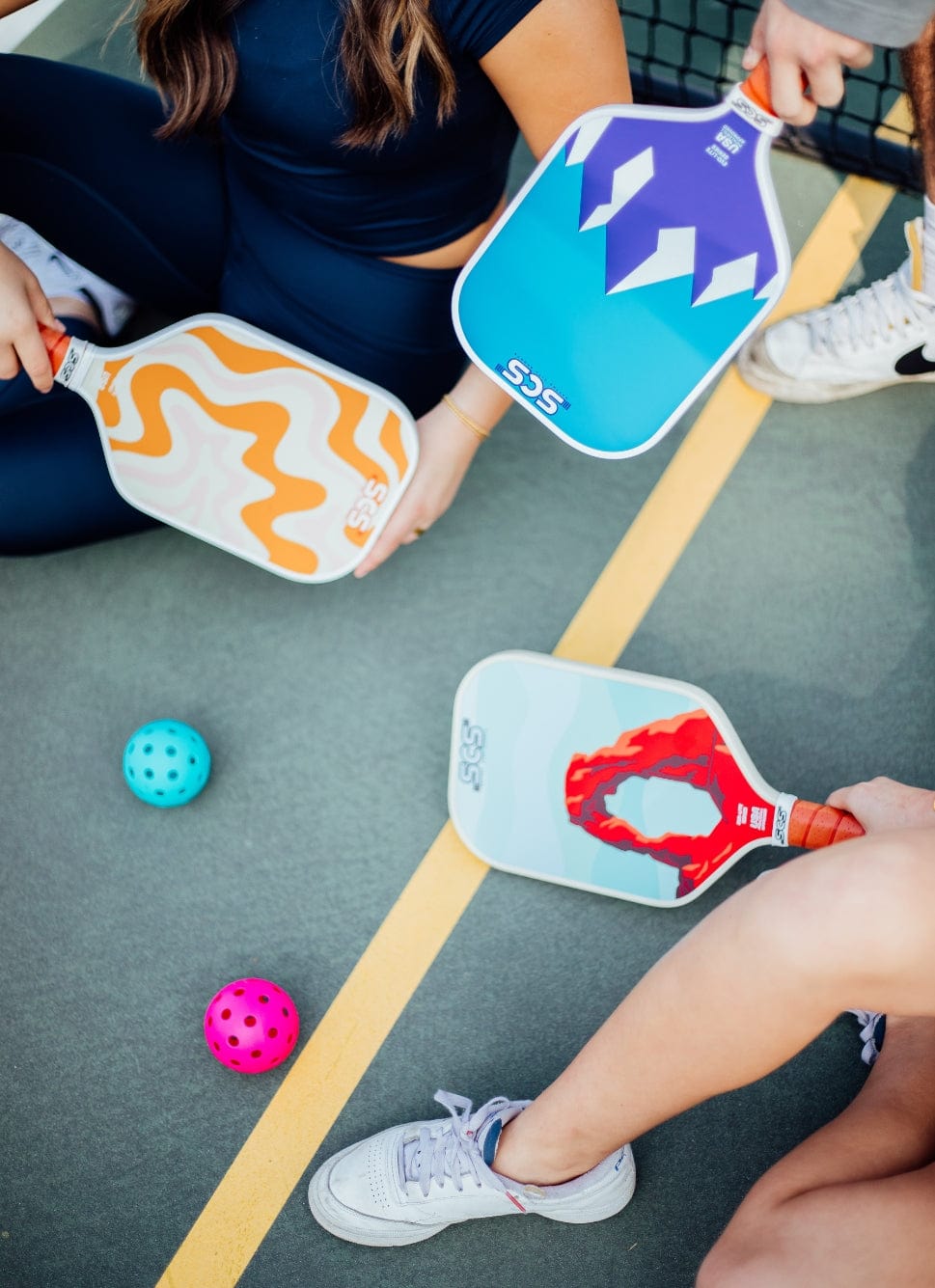 Three people holding fun and colorful pickleball paddles on a pickleball court